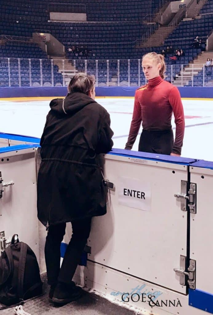 Stéphane leans at the boards of the ice rink, back to camera. He is wearing a black coat with a hood. Deniss faces Stéphane on the ice, with an intent and serious look on his face. He is wearing a red ombre turtleneck top with a gold epaulet.