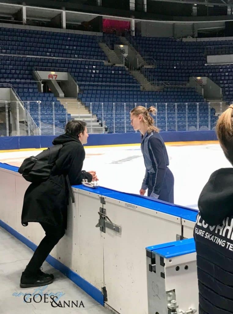 Stéphane leans at the boards of the ice rink, in profile to the camera. He is wearing a black coat with a hood, and a black backpack. Deniss is turning away from Stephane toward the center ice. He is wearing a collared shirt in two tones of grey/blue and pants of the darker color blue. His hair is half up in a bun.