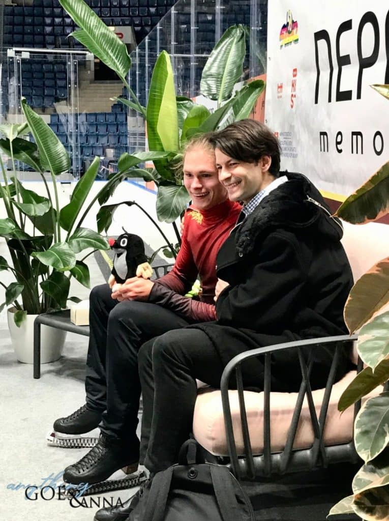 Deniss Vasiljevs and Stephane Lambiel pose in the kiss and cry at the Nepela Trophy. Deniss has a mole plushie on his knee and is wearing a red top and black pants. Stéphane is wearing a black coat and pants. Both are smiling.