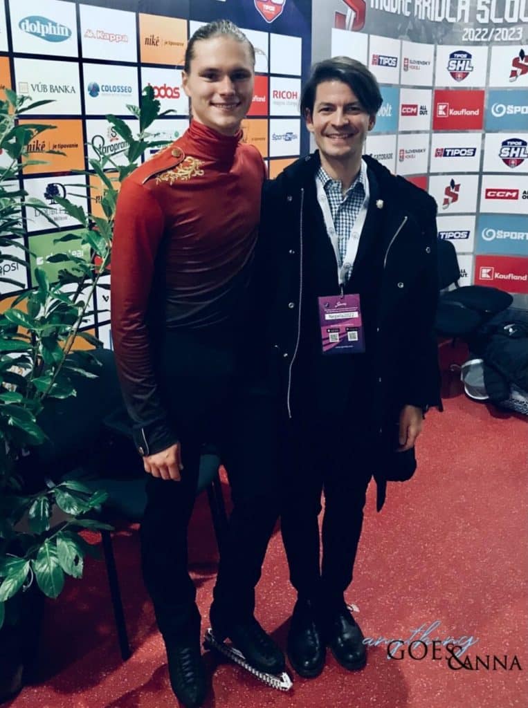 Deniss and Stéphane smile at the camera. They are standing side by side backstage, and are in front of a wall covered in sponsor logos. Deniss is wearing his free program costume and skates. Stephane is wearing a black coat, white and blue checked shirt, and black pants and black shoes.