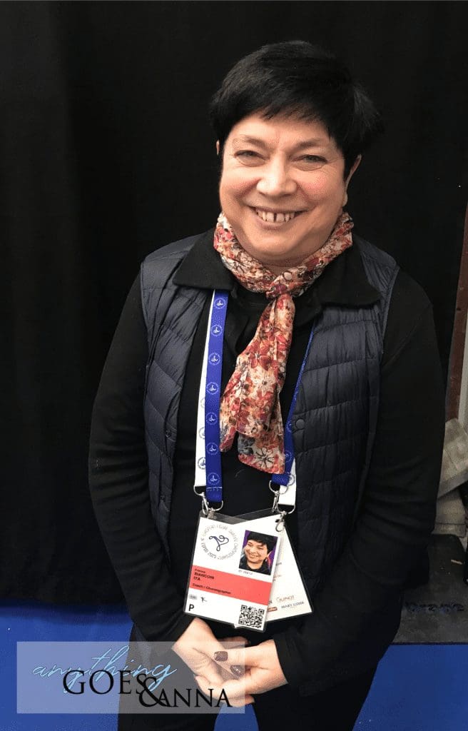 Franca Bianconi is a white woman with short black hair wearing a black shirt, black puffy vest, orange floral scarf and event badges. She is smiling at the camera.